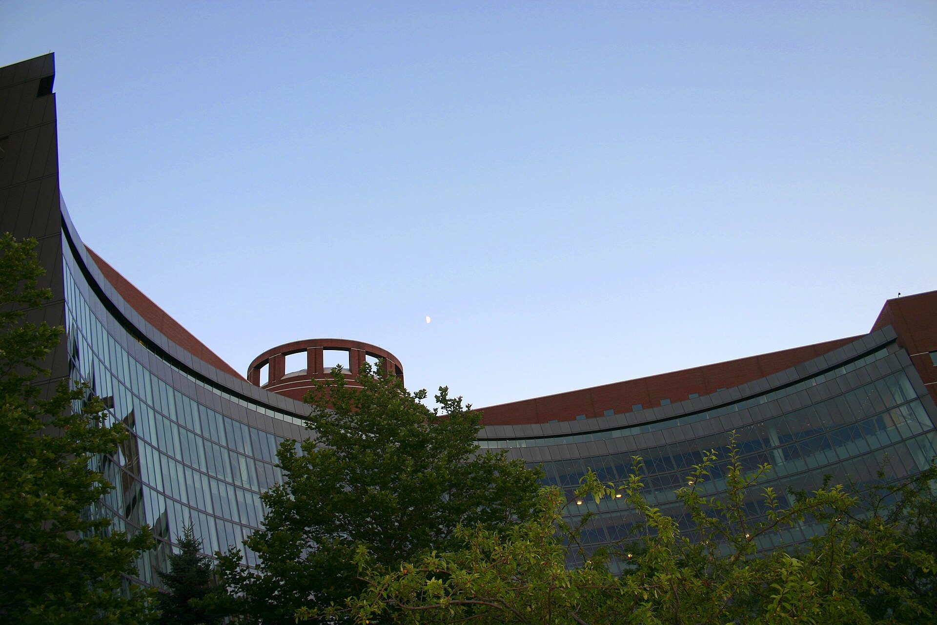 The curved glass-and-brick facade of a large federal courthouse at dusk, a round brick rotunda rising at its centre, leafy trees across the foreground and a half moon in a clear blue sky; nobody in the frame
