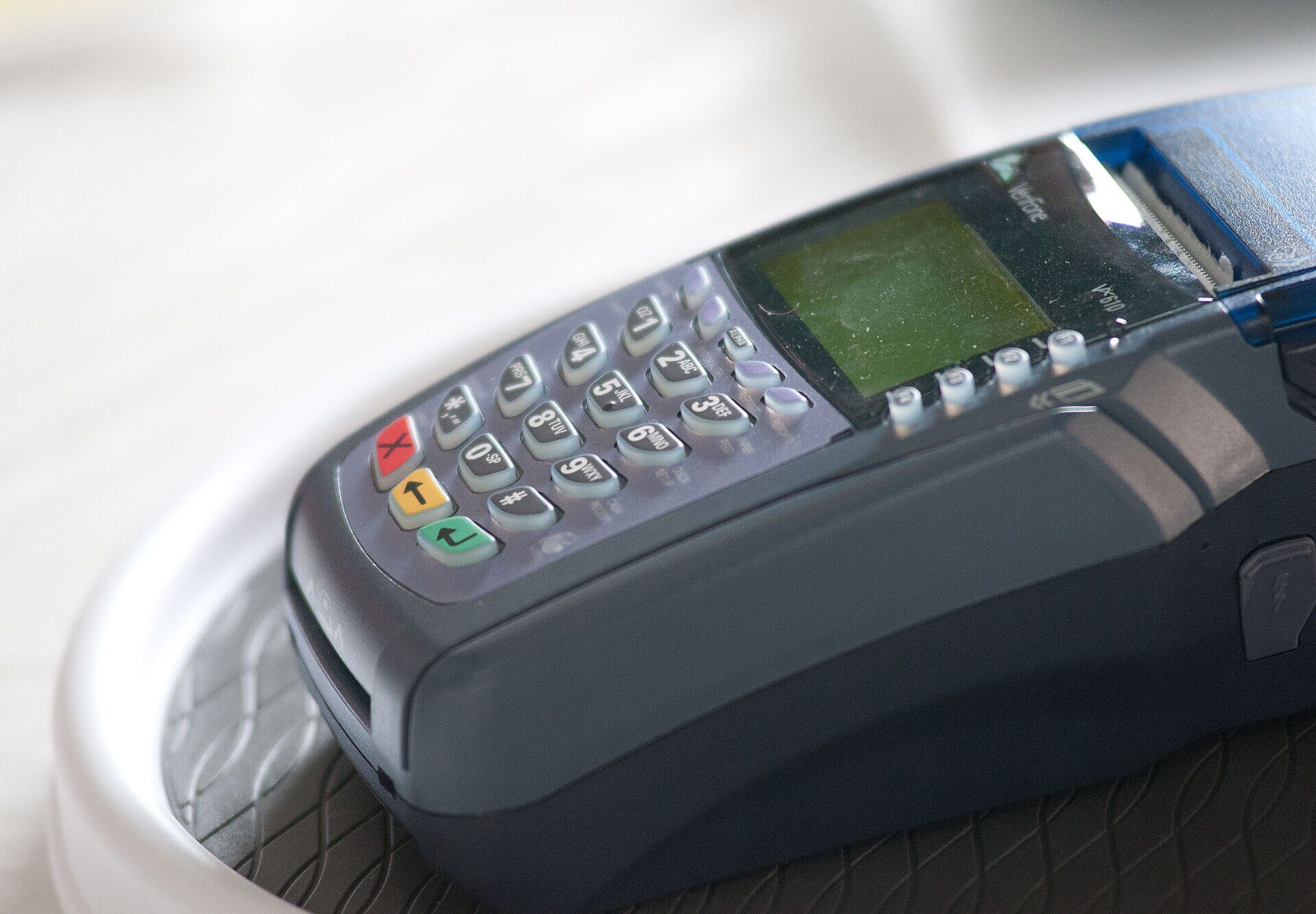 Close-up of a hand-held electronic payment terminal resting on a white table. The device has a rubber keypad with number and letter keys, a small green-grey screen, a row of function keys beneath the screen and a card slot along the front edge. No people are in the frame.