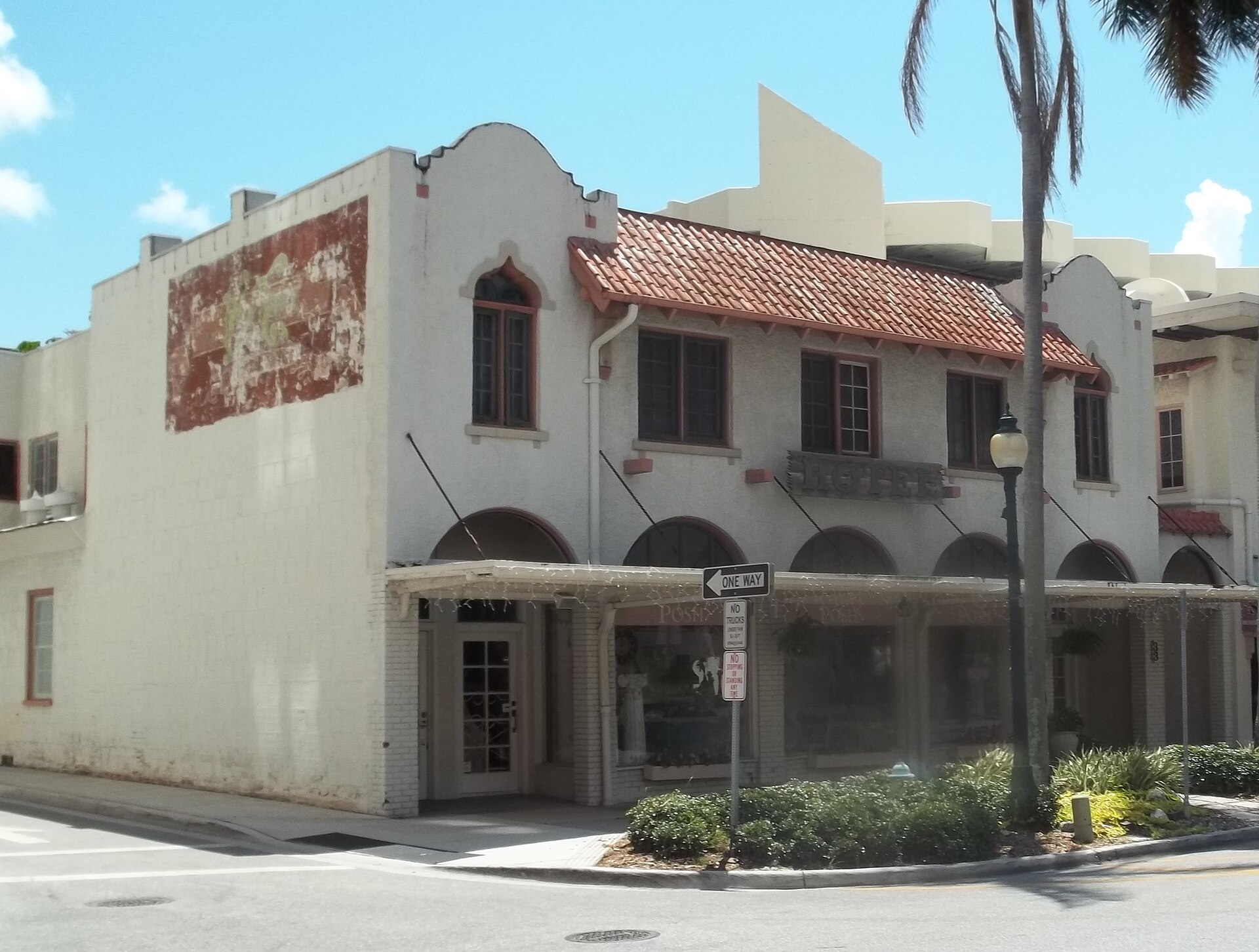 A two-storey Mediterranean Revival building on a corner in downtown Sarasota, Florida: white stucco walls, a red barrel-tile roof over an arcade of arched shop windows, arched upper windows with dark frames, metal letters spelling HOTEL across the facade, a faded painted advertisement on the blank side wall, a palm tree and a black lamp-post at the kerb. Street signs read ONE WAY, NO TRUCKS and NO STOPPING OR STANDING ANY TIME. The pavement and the street are empty; nobody is in the frame.