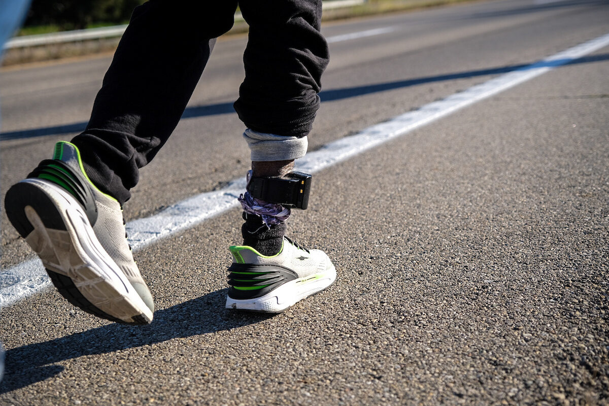 Close view of a person's lower legs in mid-stride on an asphalt road, beside a painted white line. A black GPS ankle monitor with a wide strap is fastened over the rolled cuff of dark trousers, above a dark sock and a white running shoe with green trim. The road surface fills the frame and no face is visible.