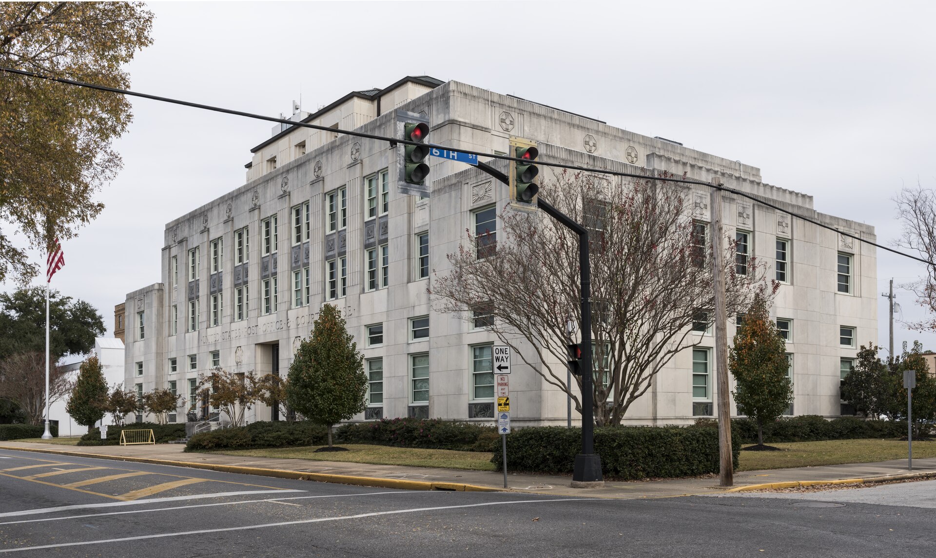 A pale stone federal building on a street corner with UNITED STATES POST OFFICE AND COURTHOUSE carved across its facade, a traffic light and a 6th Street sign in front of it, an American flag on a pole at the left, nobody in the frame