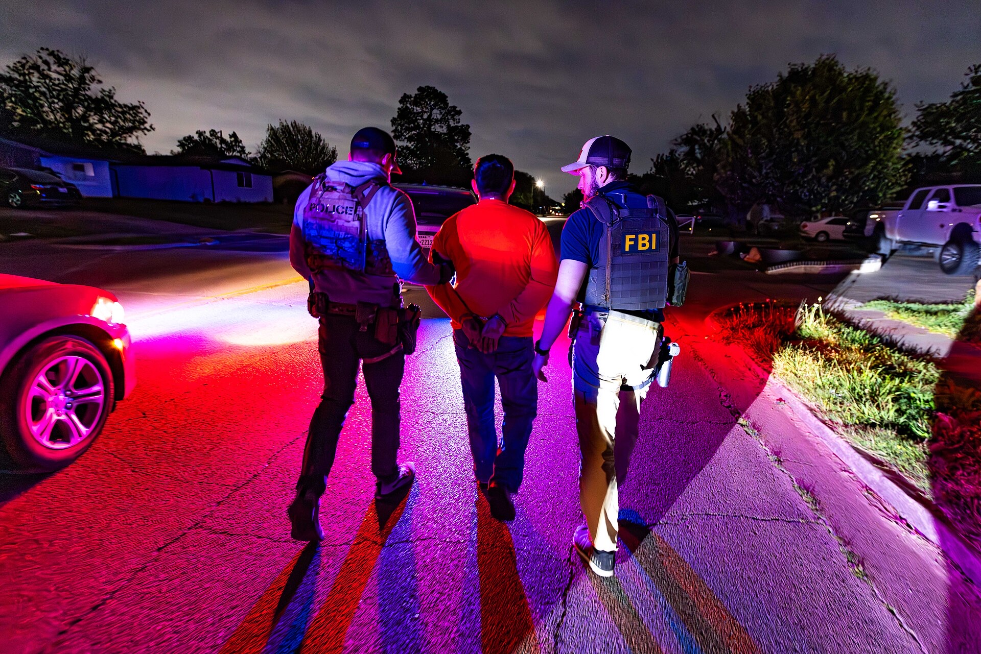 Seen from behind at night under red and blue lights, an ICE officer and an FBI agent walk a handcuffed man in an orange shirt down a residential street