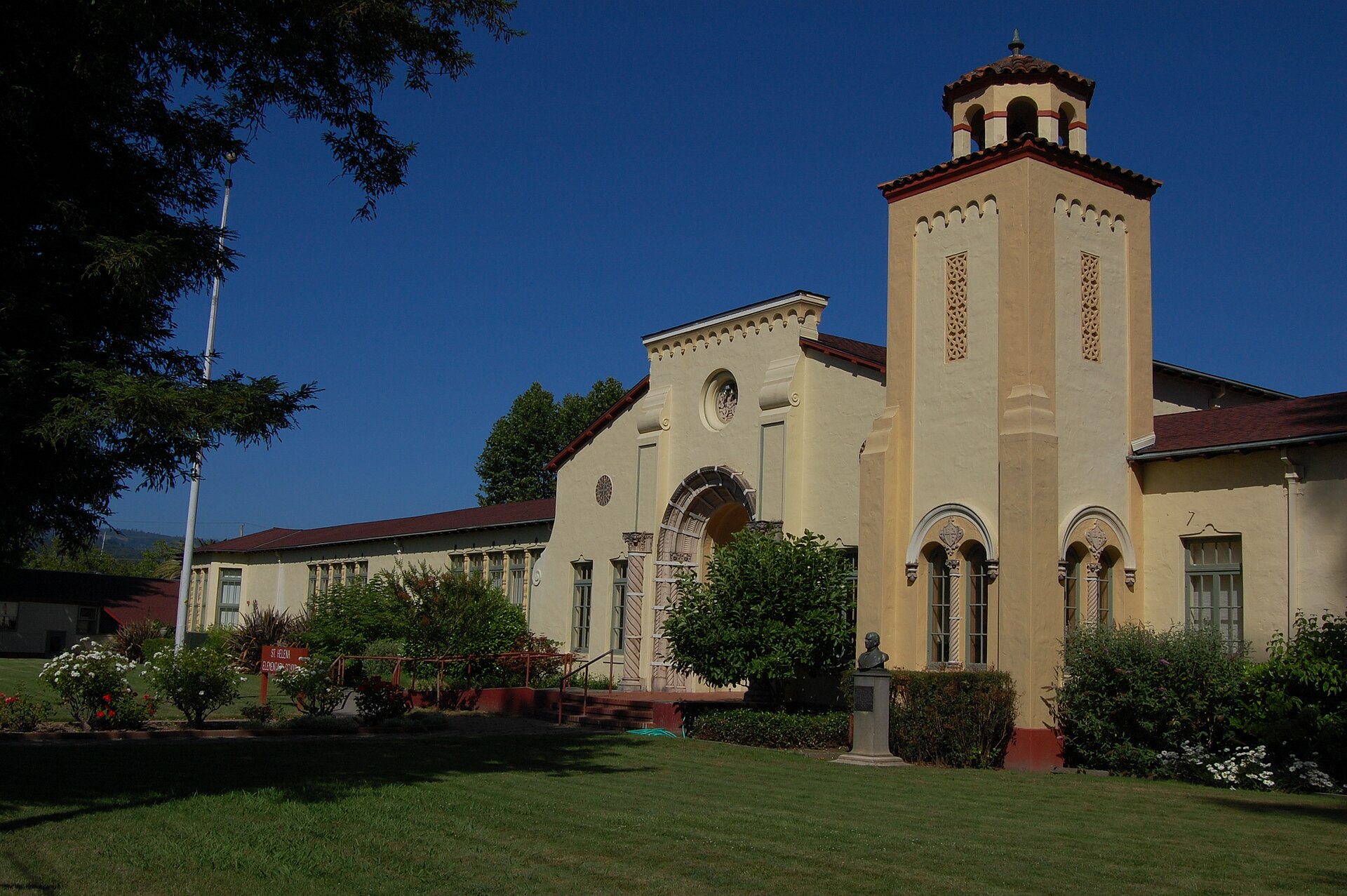 A cream-coloured elementary school with a red tile roof and a bell tower behind an empty green lawn, no people in the frame