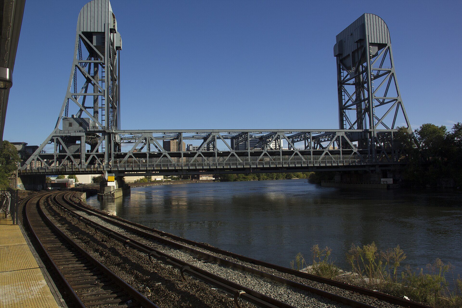 A steel lift bridge with two tall towers spans a river under a clear blue sky. Railway tracks curve along the near bank toward the bridge, with the yellow edge of a station platform at the left. Apartment buildings stand behind the bridge on the far side. There is no person in the frame.