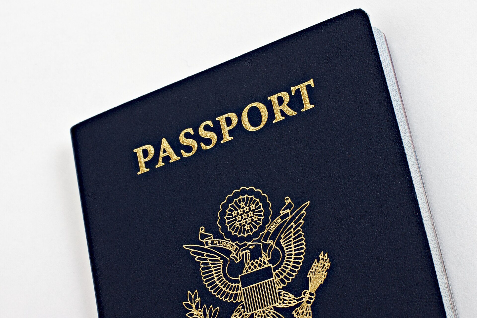 A close photograph of the cover of a United States passport, navy blue with gold lettering, lying on a white surface. The word PASSPORT is stamped across the upper part of the cover in gold capitals, and below it is the gold Great Seal of the United States: an eagle holding an olive branch and a bundle of arrows, with a scroll reading E PLURIBUS UNUM across its beak. The edges of the document's pages are visible along the right side. No person is in the frame.
