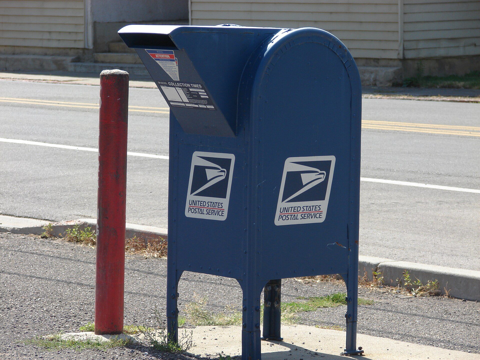 A blue United States Postal Service collection box standing on a concrete pad beside a two-lane road, photographed from the front in bright sunlight. Two USPS eagle logos and the words UNITED STATES POSTAL SERVICE are printed on its front panel, and a COLLECTION TIMES placard is fixed under the open pull-down door. A red-painted metal post stands to its left. No people are in the frame.