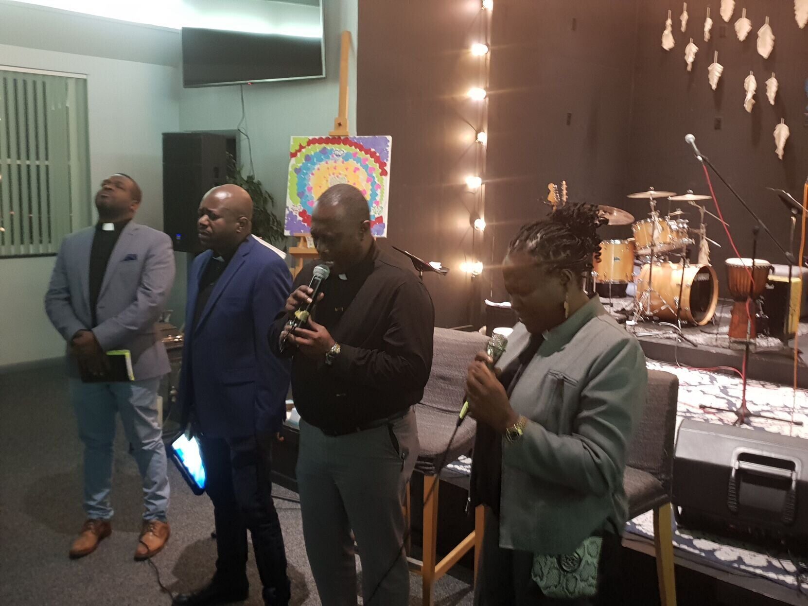 Four adults stand in a row at the front of a church hall with their heads bowed in prayer, two of them holding microphones, three wearing clerical collars. Behind them are a drum kit, a hand drum, a string of warm lights and a child's painted rainbow canvas on an easel.