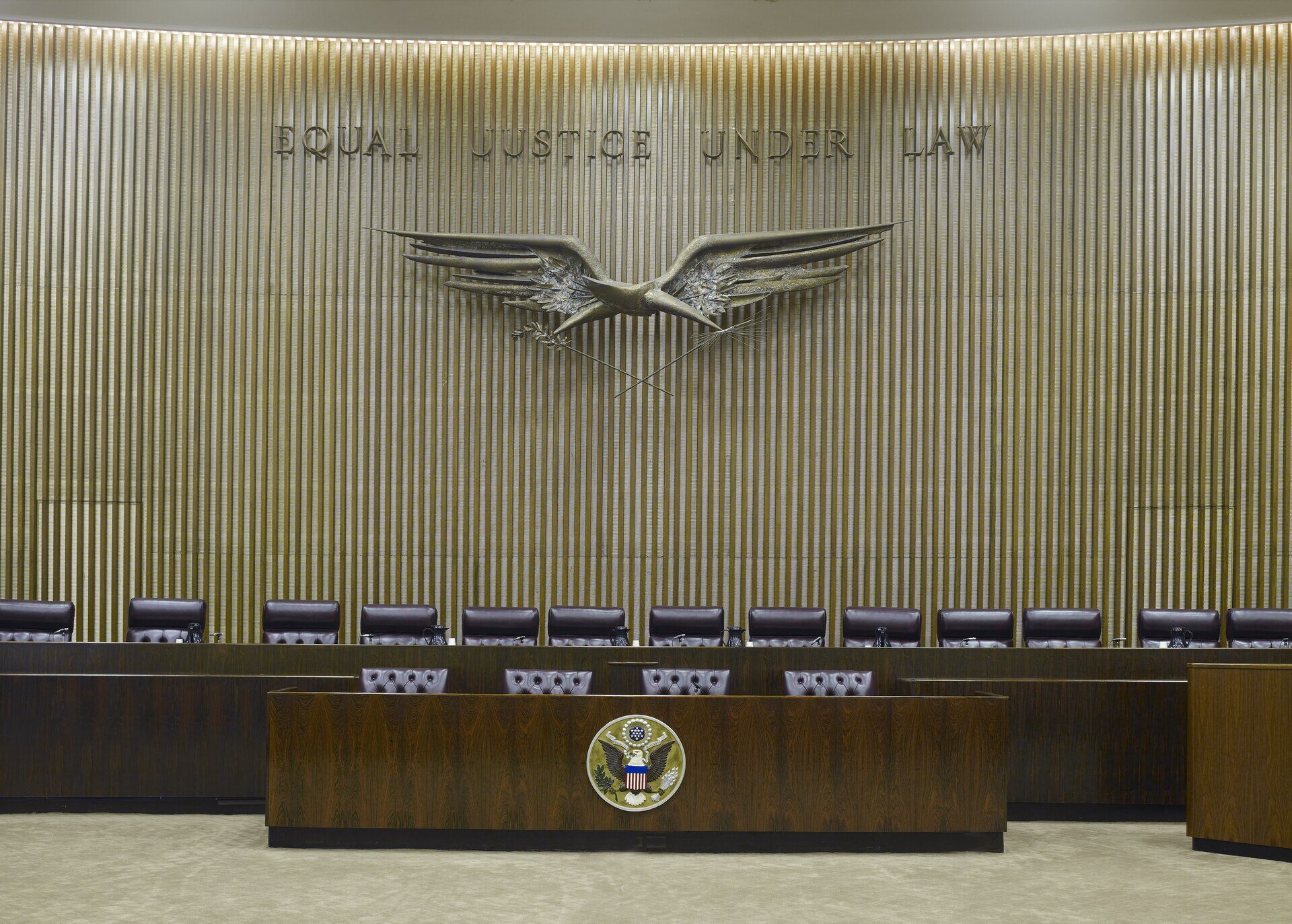 An empty courtroom photographed straight on from the floor. A curved wall of narrow vertical wooden slats fills the frame. Across the upper wall, raised metal letters read EQUAL JUSTICE UNDER LAW. Below the letters hangs a large bronze sculpture of an eagle with its wings stretched wide, holding an olive branch in one claw and a bundle of arrows in the other. In front of the wall runs a long dark wooden bench with about fifteen empty high-backed leather chairs behind it, each with a small microphone and a water jug. Lower and closer to the camera is a second wooden desk with four more empty chairs, carrying the round Great Seal of the United States at its centre. The carpet is plain and pale. There is no person in the picture.