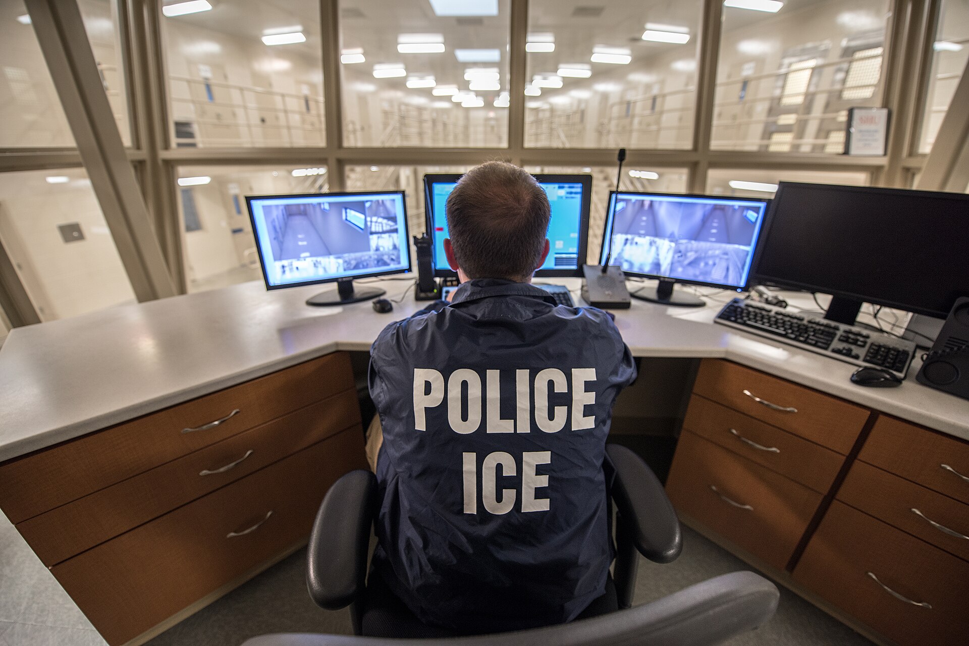 A control desk inside an immigration detention facility, photographed from behind: an officer in a dark blue vest lettered POLICE ICE across the back sits facing four monitors, three of them showing black-and-white camera views of empty corridors and cell doors, with a handheld radio and two keyboards on the counter; behind the desk, tall glass windows look out on a two-level cell block with railed walkways, numbered doors and fluorescent lighting. No detainee is visible and the officer's face cannot be seen.