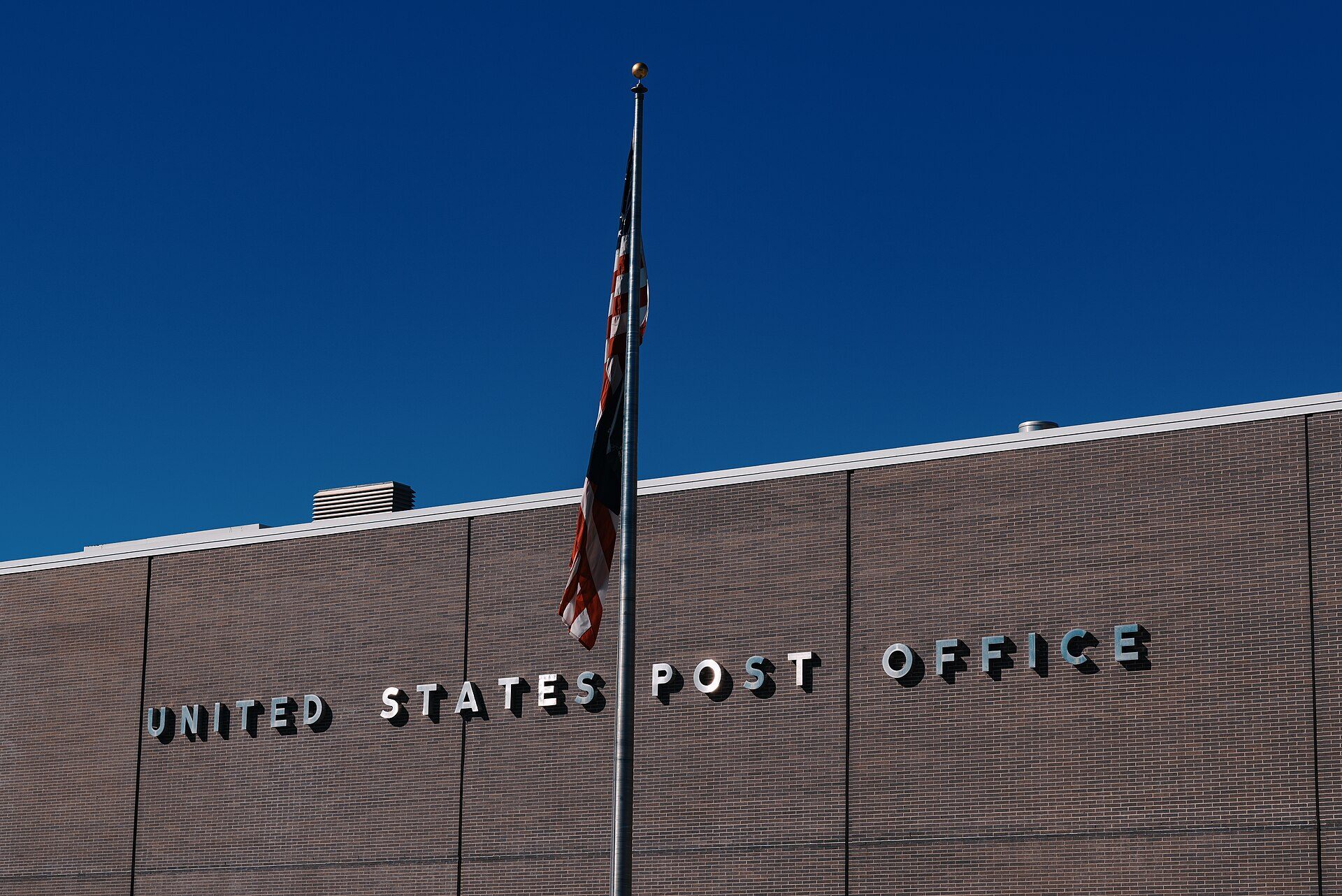 The upper facade of a brick United States post office building against a deep blue sky, photographed from below. Raised metal letters reading UNITED STATES POST OFFICE run along the brickwork. A flagpole with a furled United States flag stands in front of the building, and a roof vent is visible at the left. No people are in the frame.