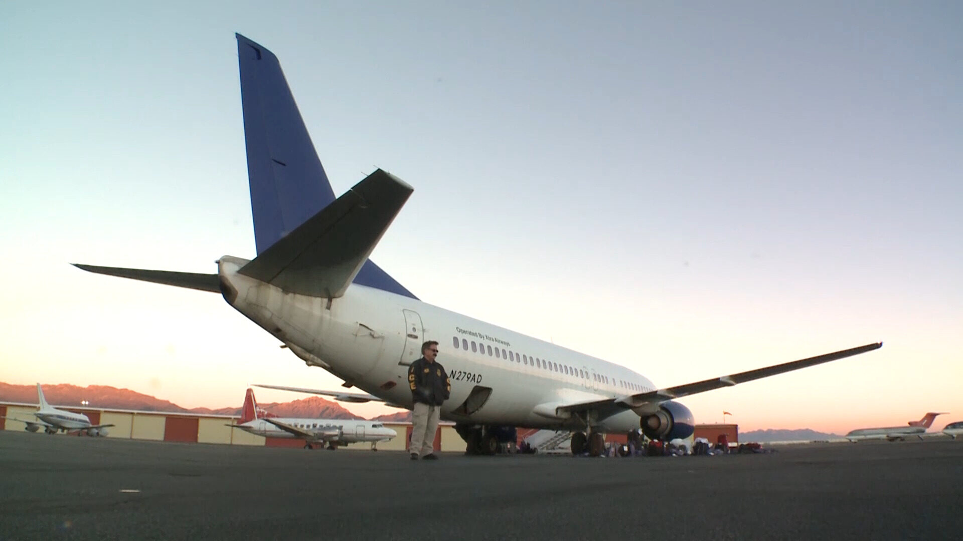A white charter jet parked on an airport apron at dawn, photographed from low and behind the tail. The registration N279AD is painted on the rear fuselage beside the words Operated By Xtra Airways. One uniformed officer in a dark jacket marked ICE stands alone beneath the tail, facing the camera at a distance. Smaller propeller aircraft and low hangars sit along the apron behind, with a mountain ridge and an orange sunrise on the horizon.