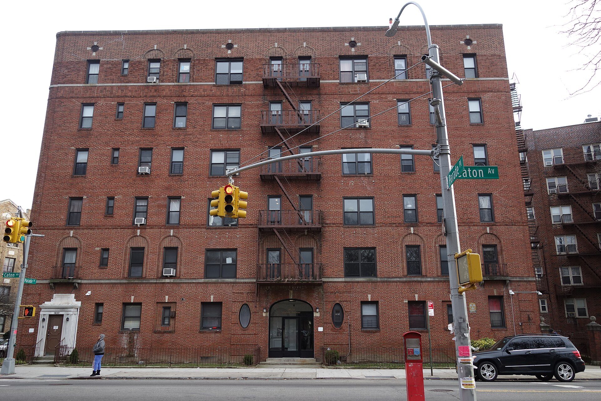 A six-storey red brick apartment building on a street corner in Brooklyn, photographed from across the road. A glass double door sits under an arched stone entrance at the centre of the ground floor, with a second, smaller doorway at the left end of the building. Fire escapes run up the middle of the facade, green street signs on the corner pole read Caton Av and Parade Pl, and a traffic light hangs over the intersection. One adult stands on the pavement near the left-hand door, turned away from the camera.