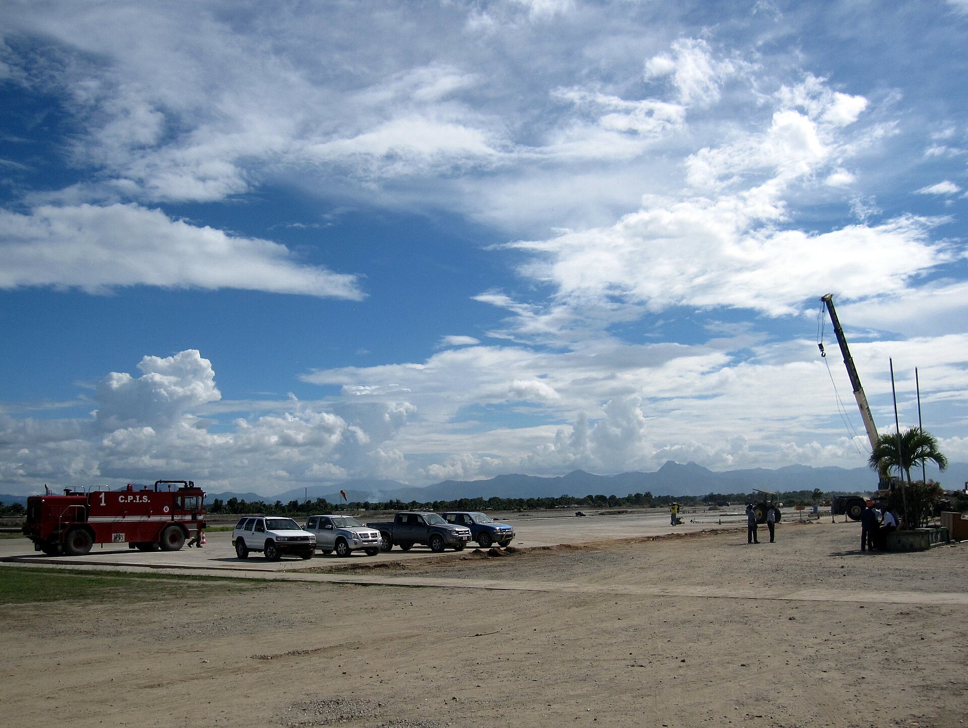 The apron at Cap-Haïtien's airport: a red fire truck, four parked vehicles, a crane, workers at a distance, mountains on the horizon under a wide sky